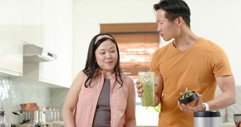 Couple making green smoothie in bright sunlit kitchen smiling while holding blender cup
