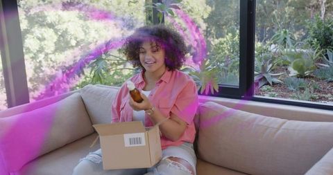 Curly woman unpacking amber bottle on sofa, smiling during casual unboxing in daylight