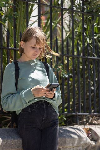 Young woman in early 20s leaning on ledge checking smartphone by iron fence in sunlight