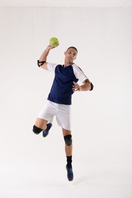 Young male athlete in mid-air throwing handball, wearing sports uniform with knee and elbow pads. Ideal for use in sports-related content, athletic training materials, competitive game promotions, and fitness advertisements.