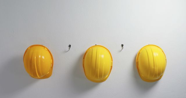 Three bright yellow safety helmets are hanging on hooks attached to a white wall. Ideal for use in industrial safety training materials, construction site promotions, workplace safety campaigns, or images depicting organized working environments. The image emphasizes protection, orderliness, and readiness in a professional setting.