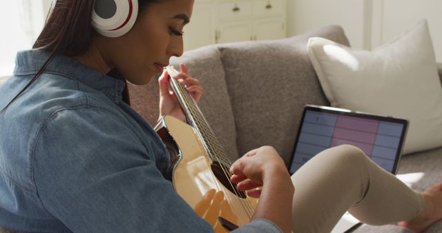 Image of happy biracial woman in headphones sitting on sofa and playing guitar. Lifestyle, music, hobby and spending free time at home concept.