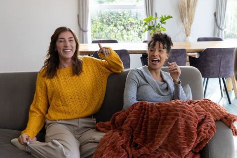 Laughing friends relaxing on sofa in mustard sweater with rust throw cozy daylight moment