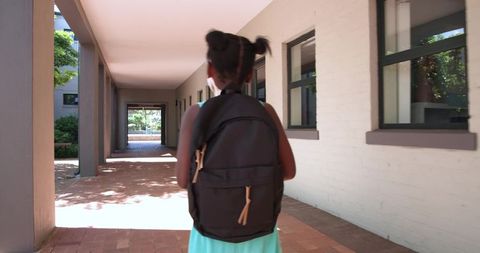 African schoolgirl walking sunlit campus corridor carrying backpack in turquoise dress