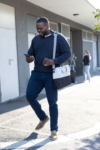 African American commuter walking with coffee and messenger bag while checking smartphone