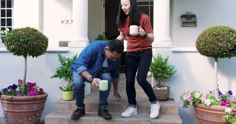 Asian couple holding ceramic mugs on tiled porch steps with potted topiary and flowers