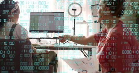 Woman pointing at monitor while collaborating on digital charts with binary data overlay