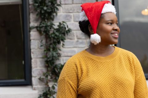African american woman wearing santa hat, smiling outdoors in festive mustard knit sweater