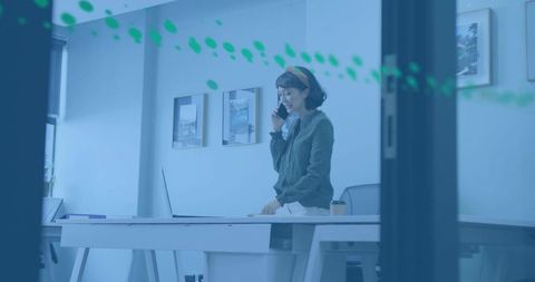Talking asian woman on phone at desk in glass office, wearing green cardigan and headband