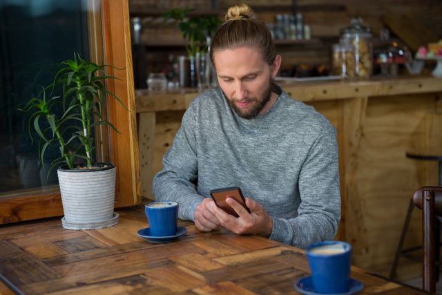 Young male hipster with beard and man bun using mobile phone at wooden table in coffee shop. Blue coffee cups and potted plant on table. Ideal for themes of modern lifestyle, technology use, casual settings, and coffee culture.