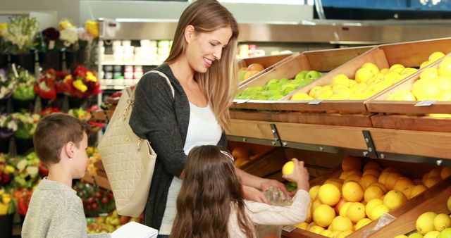 Mother shopping for fresh fruits with children at grocery store. She is helping her daughter pick out oranges from a wooden display, while her son stands close by. Perfect for illustrating family lifestyle, healthy eating habits, and grocery shopping ideas. Ideal for use in advertisements, blogs, and articles related to nutrition, parenting, and everyday life.