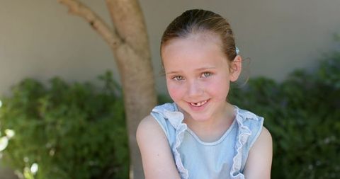 Smiling young girl in blue dress by tree, natural light backyard portrait, family lifestyle