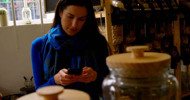 Woman wearing a blue scarf and sweater is engrossed in her smartphone in a quaint, cozy store with jars and dispensers in the background. This image can be used to promote mobile technology, online shopping apps, cozy lifestyle settings, and modern communication.