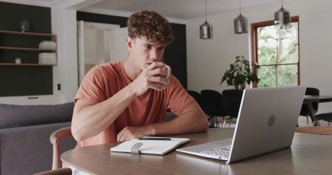 Young man working from home with laptop, coffee and notebook at modern wooden dining table