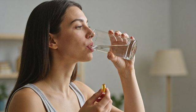 Young woman drinking water while holding yellow softgel supplement capsule at home