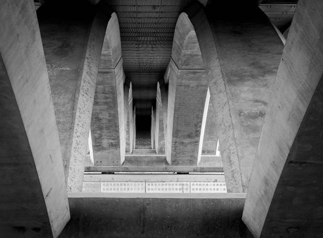 A black and white architectural view illustrating symmetry and geometry in a modern concrete bridge's arches. Elevating sense of urban infrastructure and engineering, this can be used in projects related to architecture, structural engineering, or design. Perfect for backgrounds, web design, urban exploration blogs, and architectural studies.