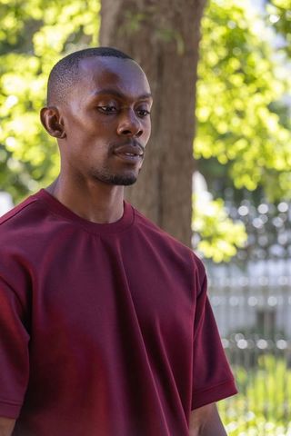 Contemplative african american man wearing maroon t-shirt standing in sunlit park