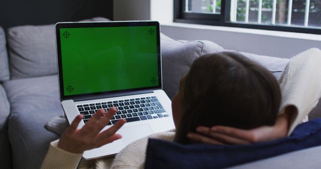 This image shows someone relaxing on a gray couch with a laptop on their lap, displaying a green screen. Perfect for illustrating themes of leisure, remote work, technology at home, or comfort. Useful for articles or blog posts about working from home, living room setups, or relaxing with technology.