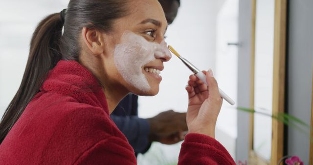 Smiling woman in a red robe applying a face mask in the bathroom. Useful for beauty and skincare campaigns, health and wellness advertisements, lifestyle blogs, and self-care guides.