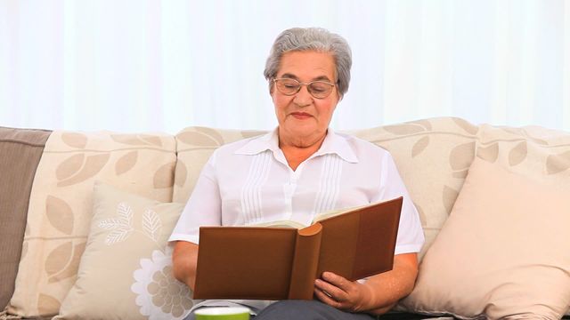 Elderly woman with gray hair and glasses is engrossed in her photo album while sitting on a patterned couch at home. This can be used to represent themes like family memories, nostalgia, relaxed lifestyle, and leisure activities for seniors. It is suitable for promotions related to senior living, family heritage, sentimental content, and elderly care.