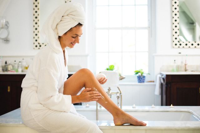 Young woman in a white bathrobe sitting on the edge of a bathtub, applying moisturizer to her leg. Ideal for use in articles or advertisements related to skincare, self-care routines, beauty products, and home wellness. Can be used to promote bath and body products, spa services, or lifestyle blogs focusing on personal care and relaxation.