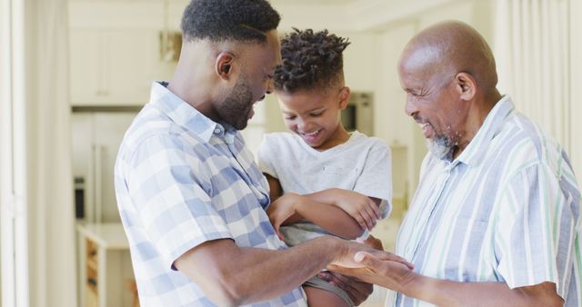 Three generations of African American men enjoying quality time at home. The image depicts a father holding his son while the grandfather interacts with them, all smiling and engaged in a warm family moment. Perfect for use in advertisements promoting family values, home life, intergenerational relationships, or Father's Day celebrations.