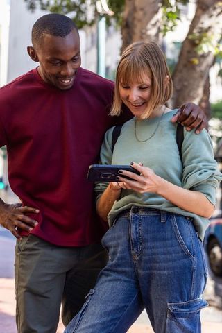 Smiling diverse couple sharing smartphone on tree-lined city sidewalk