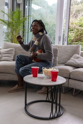 African american man cheering on couch holding football, popcorn bowl and red cups