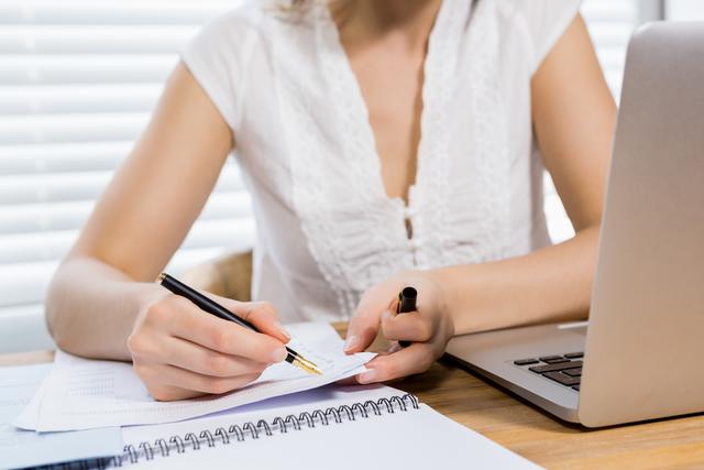 Woman writing notes on paper at home office desk with laptop. Ideal for illustrating remote work, freelance lifestyle, productivity, planning, business tasks, and home office setups.