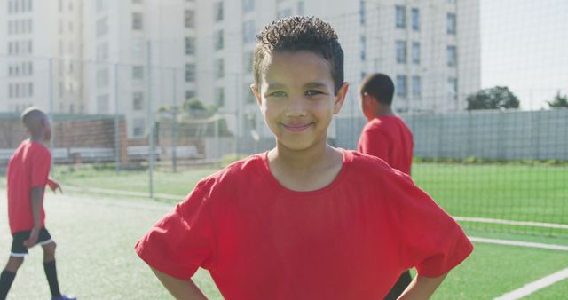 Young boy smiling confidently while standing on soccer field during a sunny day. Boy wears a red jersey, indicating he is part of a sports team. Other children in matching red jerseys are in the background. Perfect for use in articles or promotions related to youth sports, teamwork, soccer, outdoor activities, and promoting confidence in children.