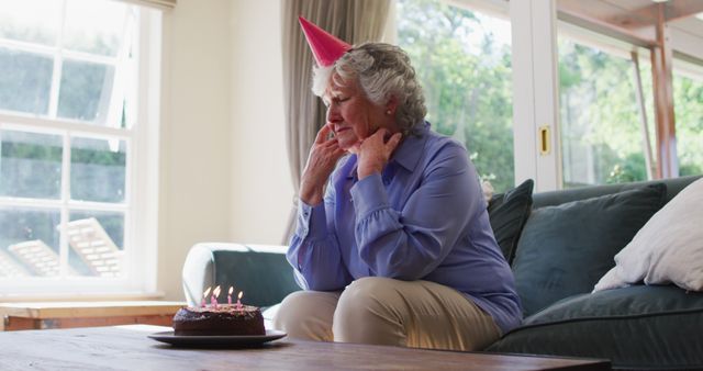 Sad caucasian senior woman with birthday cake looking out of the window at home. social distancing during coronavirus quarantine lockdown.