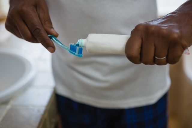 This image shows a man applying toothpaste on a toothbrush in a bathroom. It is ideal for use in articles or advertisements related to dental hygiene, personal care routines, or health and wellness tips. The close-up focus on the hands and toothpaste emphasizes the importance of daily dental care.