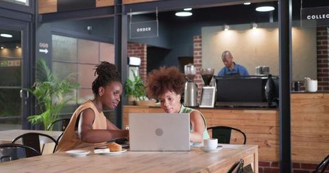 Two women collaborating on laptop in modern industrial cafe with coffee and croissant