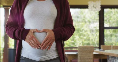Pregnant woman making heart shape with hands on belly in home interior