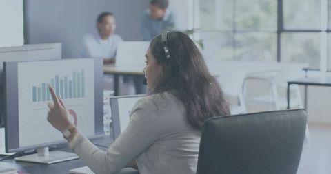 Female analyst presenting data on monitor, pointing and wearing headset in open office
