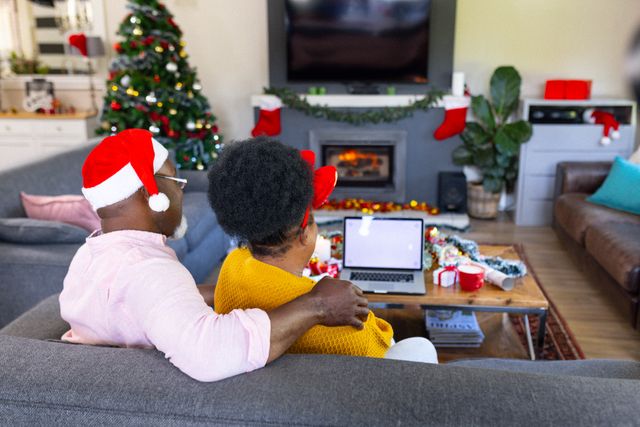 Senior African American couple sitting on sofa in living room, wearing Santa hats, using laptop with blank screen. Christmas decorations, tree, and fireplace in background. Ideal for holiday, family, and technology themes, showcasing festive home environment and togetherness.