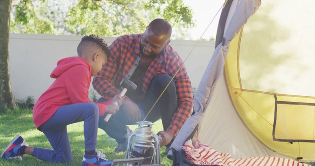 Father and son are actively engaged in setting up a tent in their backyard, with both focusing on their respective tasks: father holding stakes, son holding a hammer. Ideal for themes of family bonding, outdoor activities, parenting moments, and teamwork.