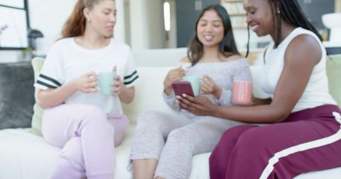 Diverse women sharing smartphone and mugs on sunlit sofa in cozy loungewear