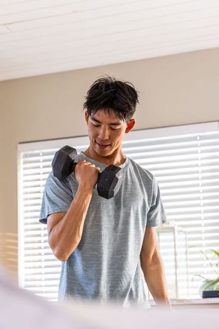 Asian man doing bicep curl with hex dumbbell in bright home gym near window blinds