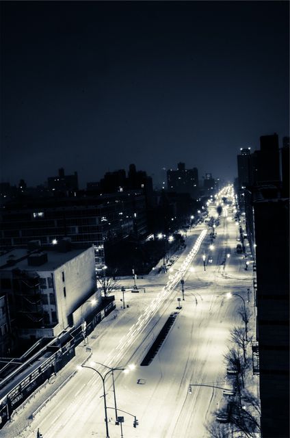 Snow-covered urban street at night featuring glowing streetlights and surrounding high-rise buildings. Ideal for themes of winter, urban living, nighttime tranquility, or cityscapes.