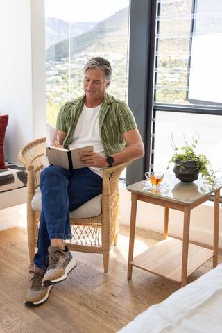 Senior man journaling in sunlit rattan chair by window with tea and mountain view