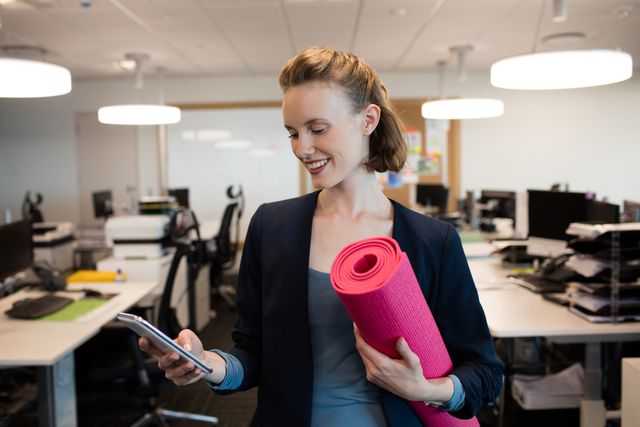Smiling businesswoman with exercise mat using mobile phone at office