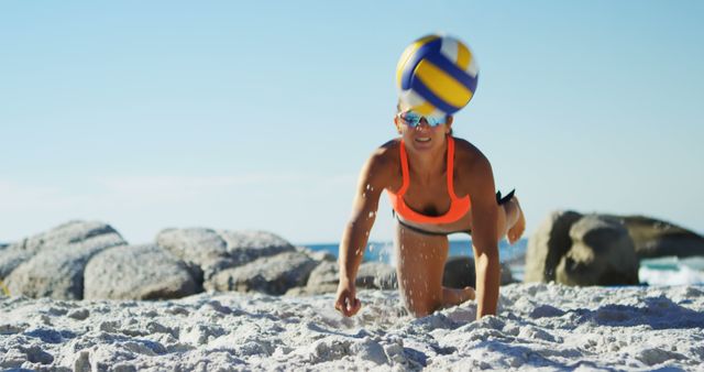 Woman playing beach volleyball, diving for the ball with intense focus, surrounded by sandy beach and clear sky. Perfect for promoting sports events, fitness programs, summer activities, and outdoor recreation.