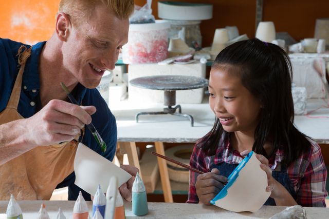 Male potter teaching young girl painting techniques in pottery workshop. Image features them smiling and enjoying the creative process. Useful for educational content, creative workshop promotions, and articles on arts and crafts.