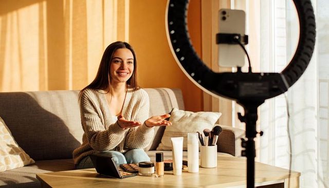 Vlogger demonstrating makeup palette with ring light on living room sofa in soft daylight
