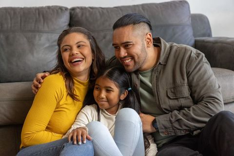 Asian family cuddling on gray sofa, parents embracing daughter, cozy home togetherness