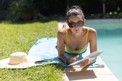 Woman reading paperback poolside on blue towel in yellow swimsuit and sunglasses