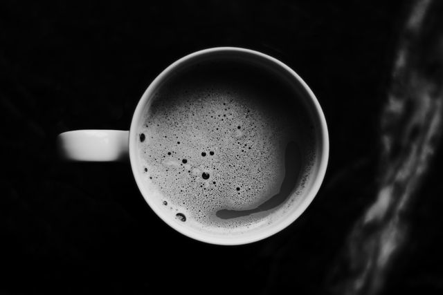 Top-down view of white coffee cup with hot frothy black coffee on dark surface. Ideal for use in food and drink advertising, website headers for coffee shops, or as part of marketing materials for breakfast or coffee products.