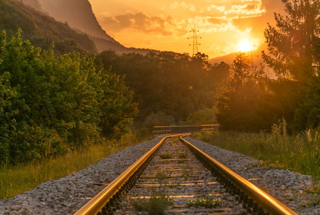 Capture of serene railroad tracks during sunset in a mountainous landscape. Ideal for themes of travel, adventure, peacefulness, nature appreciation, and environmental beauty. The golden hour lighting enhances the tranquil atmosphere, making it perfect for illustrating scenic journeys and explorations.