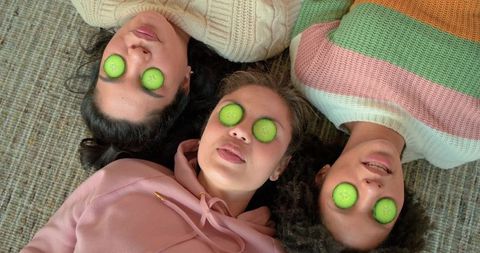 Multicultural women relaxing with cucumber eye slices on cozy carpet in pastel knits